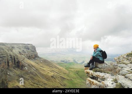 Young woman with a backpack pensively sitting on the edge of a rock and looking at the sky with clouds Stock Photo