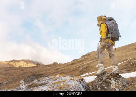 The backpacker girl in sunglasses and a big northern fur hat with a backpack on her back is standing on a rock and looking toward the cliffs hiding in Stock Photo