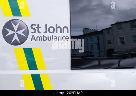 St John Ambulance logo with Maltese Cross on side of white vehicle, reflection of street in tinted window. Ballycastle, UK - August 24th, 2024. Stock Photo