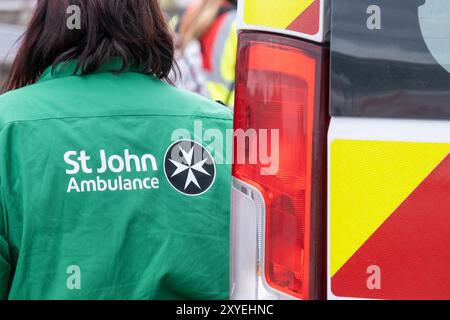 St. John's ambulance volunteer in green shirt with white lettering and black Maltese cross logo beside vehicle. Ballycastle, UK - August 26th, 2024. Stock Photo