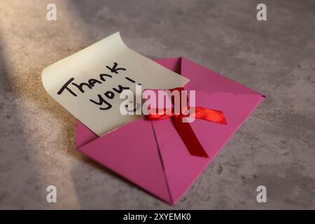 A pink envelope with a red ribbon and a note that says 'Thank You!' on it. The envelope is open and the note is written on a piece of paper. Concept o Stock Photo