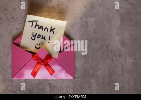 A pink envelope with a red ribbon and a note that says "Thank You!" on it. The envelope is open and the note is written on a piece of paper. Concept o Stock Photo