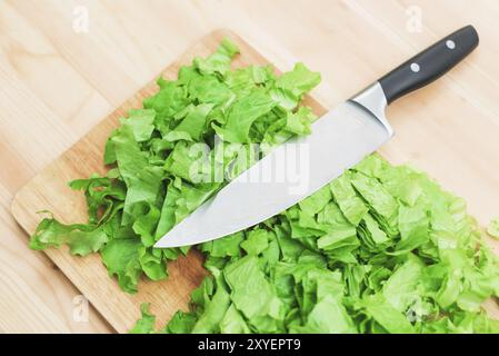 Close-up on a wooden table is a wooden cutting board on which lies the chopped lettuce leaves and on them lies a large cutting knife. The concept of s Stock Photo