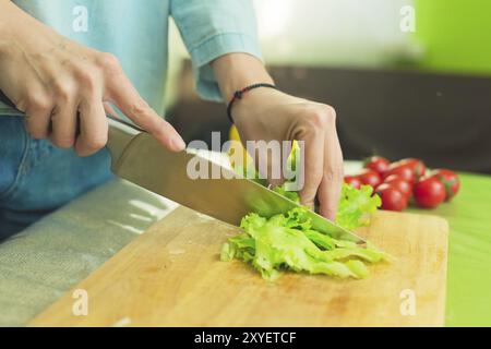 Hands of a young girl slice green lettuce leaves on a wooden cutting board on a green table in a home setting against a background of red cherry tomat Stock Photo