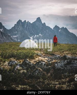 A lone hiker in a red jacket standing near a tent in a rocky, mountainous terrain with an overcast sky, in Tombstone Territorial Park in the Yukon, Ca Stock Photo