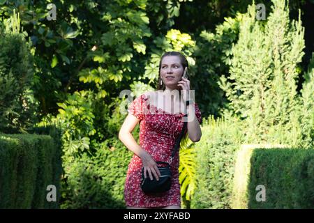 A young woman with long brown hair, wearing a red floral dress and a black bag, is having a phone conversation while walking in a lush green park on a Stock Photo