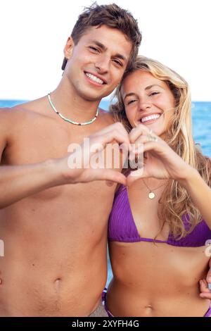 Young couple making heart shape with their hands on the beach looking at camera Stock Photo