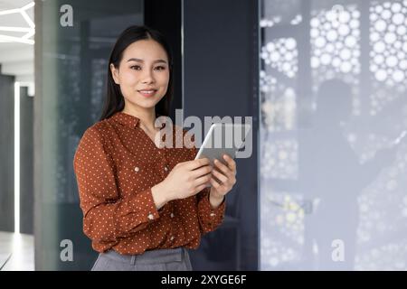 Asian worker stands confidently in modern office with tablet. Demonstrates engagement with technology. Represents digital communication and successful business applications. Stock Photo