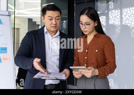 Asian business professionals engaged in discussion, focusing on project details. Man pointing to clipboard while woman with glasses holds tablet. Collaborative exchange conveys teamwork Stock Photo