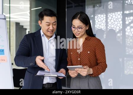 Asian business professionals engaged in collaborative discussion in modern office environment. Man and woman review documents and tablet, demonstrating teamwork and technology use. Stock Photo