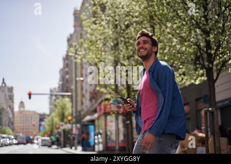 A man is smiling and holding a cell phone while standing on a city street. The scene is bright and sunny, and the man is enjoying his time outdoors Stock Photo