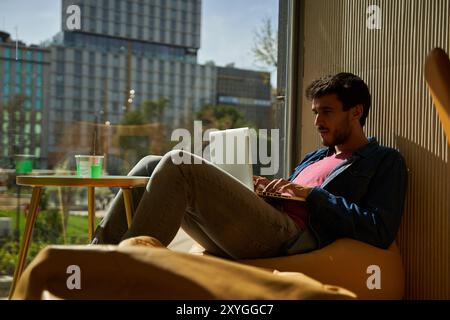 A young male student in a casual clothes resting in a comfortable pouf while studying makes notes in a laptop, freelancer works remotely Stock Photo