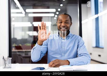 Smiling professional waves hand during video call in modern office. Engaging in virtual communication , creating friendly atmosphere. Represents business, communication, teamwork, technology Stock Photo