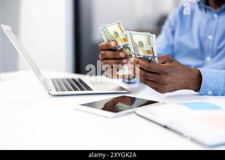 Businessman counting cash at office desk with laptop and phone. Image reflects finance, technology, accounting, and money management in professional setting. Stock Photo