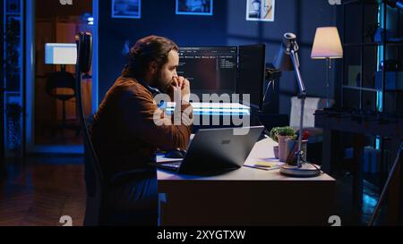 Technician focused on typing complex code in neon lit home office, developing software application. It specialist removing headphones to better concentrate on difficult task, camera B Stock Photo