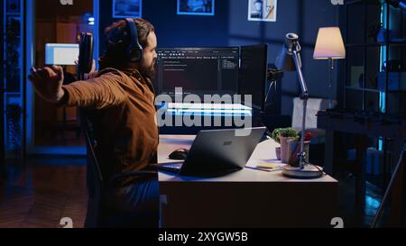 Software programmer taking break from coding, putting headphones on and stretching on desk chair during remote job shift. IT professional pausing tasks, listening music, loosening up, camera B Stock Photo