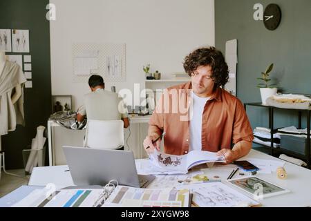 Fashionable designer turning over album pages with clothing sketches while tailor sitting at table and using sewing machine in background Stock Photo