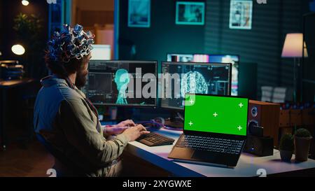 IT admin coding and using EEG headset to upload brain into green screen laptop, gaining immortality. Neuroscientist engineering transhumanism experiment on chroma key device, camera B Stock Photo