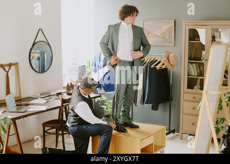 Tailor standing on one knee and doing measurements while curly haired model looking in mirror Stock Photo