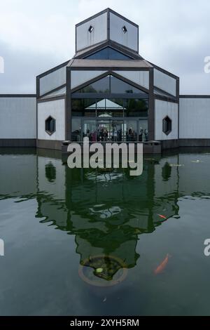 Suzhou, China - June 12, 2024 : A modern building with a large glass entrance reflects in a still pond with a few lily pads and orange fish swimming. Stock Photo