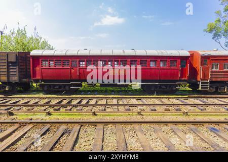 Side view of an old stationary red passenger carriage car sitting parked on train tracks, part of Sri Lanka Railways, on a blue sky day. Horizontal Stock Photo