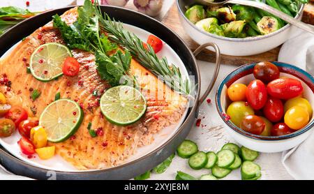 Grilled salmon and vegetables on wooden light background. Dinner table with cooked fish, tomatoes, asparagus. Stock Photo