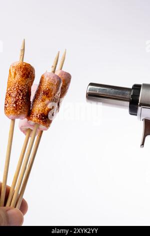 Horizontal image showing toasted marshmallows on skewers being roasted with a torch. The hand holding the torch is on the left, set against a clean wh Stock Photo