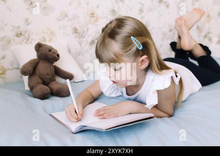 A little girl lies on the bed in the bedroom and writes in a blue book. Education concept. Home schooling. Homework. Stock Photo