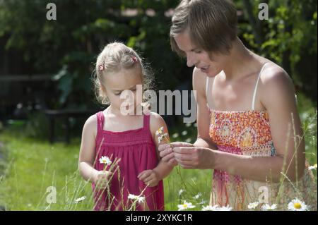 Young mother and her little daughter picking daisies on a meadow in summer to weave a wreath of flowers. Mother with her little daughter picking flowe Stock Photo