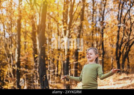 Small happy smiling little caucasian child girl playing with yellow leaves in autumn forest or garden around trees. Kid throws leaves up and they are Stock Photo