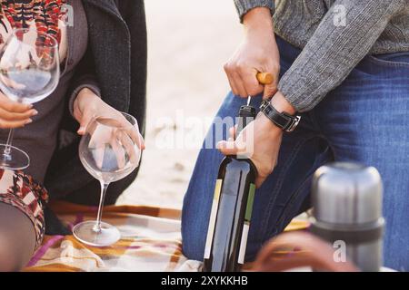 Young happy couple enjoying picnic on the beach together. Man opening bottle of wine. Close up Stock Photo