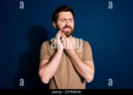 Photo of unhappy man with stylish beard dressed beige t-shirt hold hand on cheek have toothache isolated on dark blue color background Stock Photo