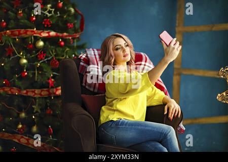 Young pretty woman taking selfie at home while sitting on armchair near Christmas tree Stock Photo