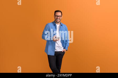 Portrait of smiling businessman with hand in pocket texting on smart phone over orange background Stock Photo