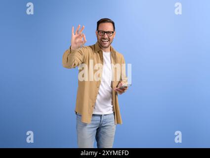 Cheerful handsome entrepreneur holding smart phone and gesturing OK sign on isolated blue background Stock Photo