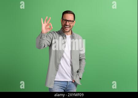 Portrait of handsome freelancer showing OK sign and laughing ecstatically against green background Stock Photo