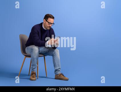Focused serious businessman checking social media apps over smart phone on chair on blue background Stock Photo