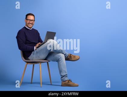 Portrait of male professional smiling and using wireless computer on chair against blue background Stock Photo