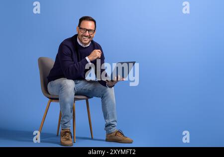 Successful male manager with laptop smiling at camera while sitting on chair over blue background Stock Photo