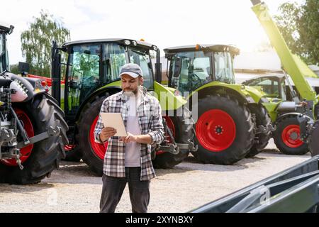 Agribusiness. Agricultural and construction equipment dealer. Man stands against new tractors and using digital tablet. Stock Photo