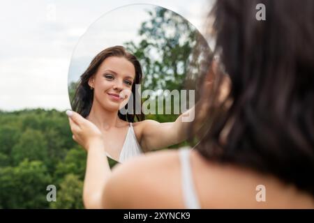 A lady holds a round mirror, reflecting her face amidst a green natural backdrop. Image contains noise and motion blur Stock Photo