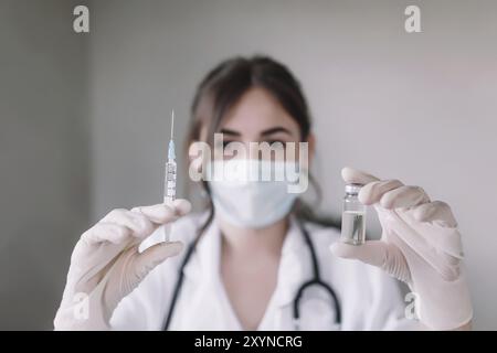Doctor or nurse in protective face mask and gloves with stethoscope is holding glass vial bottle of anti virus vaccine and syringe. Stock Photo
