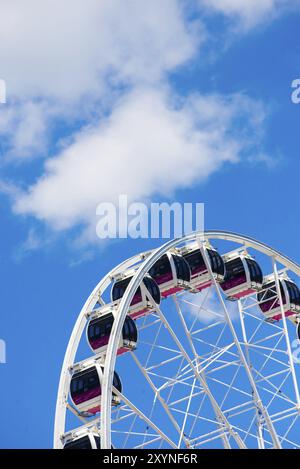 The pods of a Ferris wheel set infront of a backdrop of blue sky and white clouds, viewed from below Stock Photo