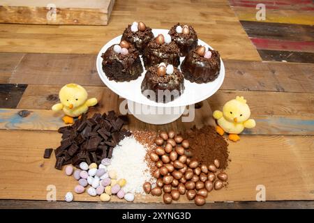 A selection of Easter Chocolate nest Cakes topped with three little chocolate mini eggs on a white plate surrounded by the ingredients and two yellow Stock Photo