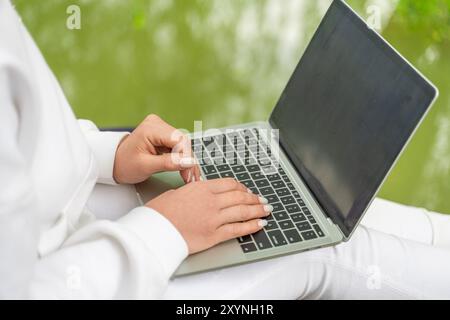 A woman is typing on a laptop in a park. The laptop is open and the screen is black. The woman is wearing a white shirt and is sitting on a bench Stock Photo