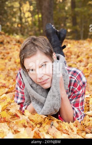 Attractive woman lying on her stomach in the beautiful autumn leaves of a maple tree. Fashionable, robust outdoor clothing. Symbolising the changing o Stock Photo