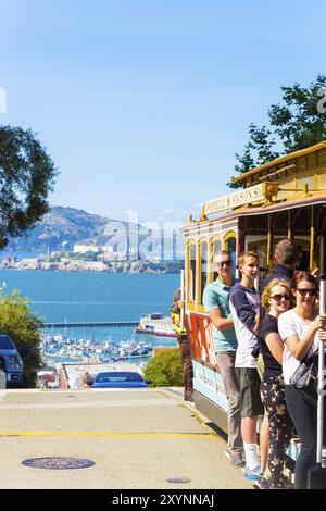 San Francisco, USA, May 15, 2016: Closeup of passengers standing on outside platform hanging on to handrails of iconic cable car with view of Alcatraz Stock Photo