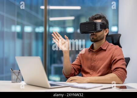 Young professional in modern office using virtual reality headset while interacting with laptop for innovative experience. Person explores new technology concepts, enhancing digital interaction Stock Photo