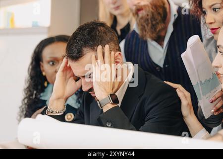 Businessman is suffering from headache at work, touching his head with hands, while his team is trying to show him some positive results Stock Photo
