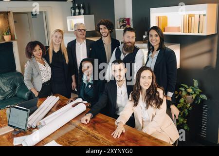 Diverse team of architects and engineers are gathered around a table in their office, smiling Stock Photo